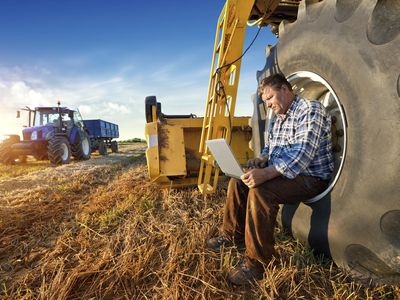 Farmer resting on a tractor wheel while working on a laptop