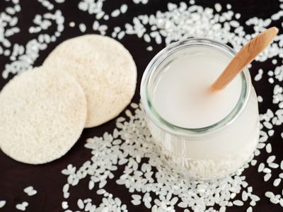 Glass jar of rice water with a wooden spoon and rice grains scattered on the table