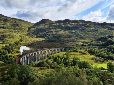 Aerial view of the lush green hills of the Scottish Highlands over the Glenfinnan viaduct with the Jacobite Steam Train passing over it under a blue sky with white clouds