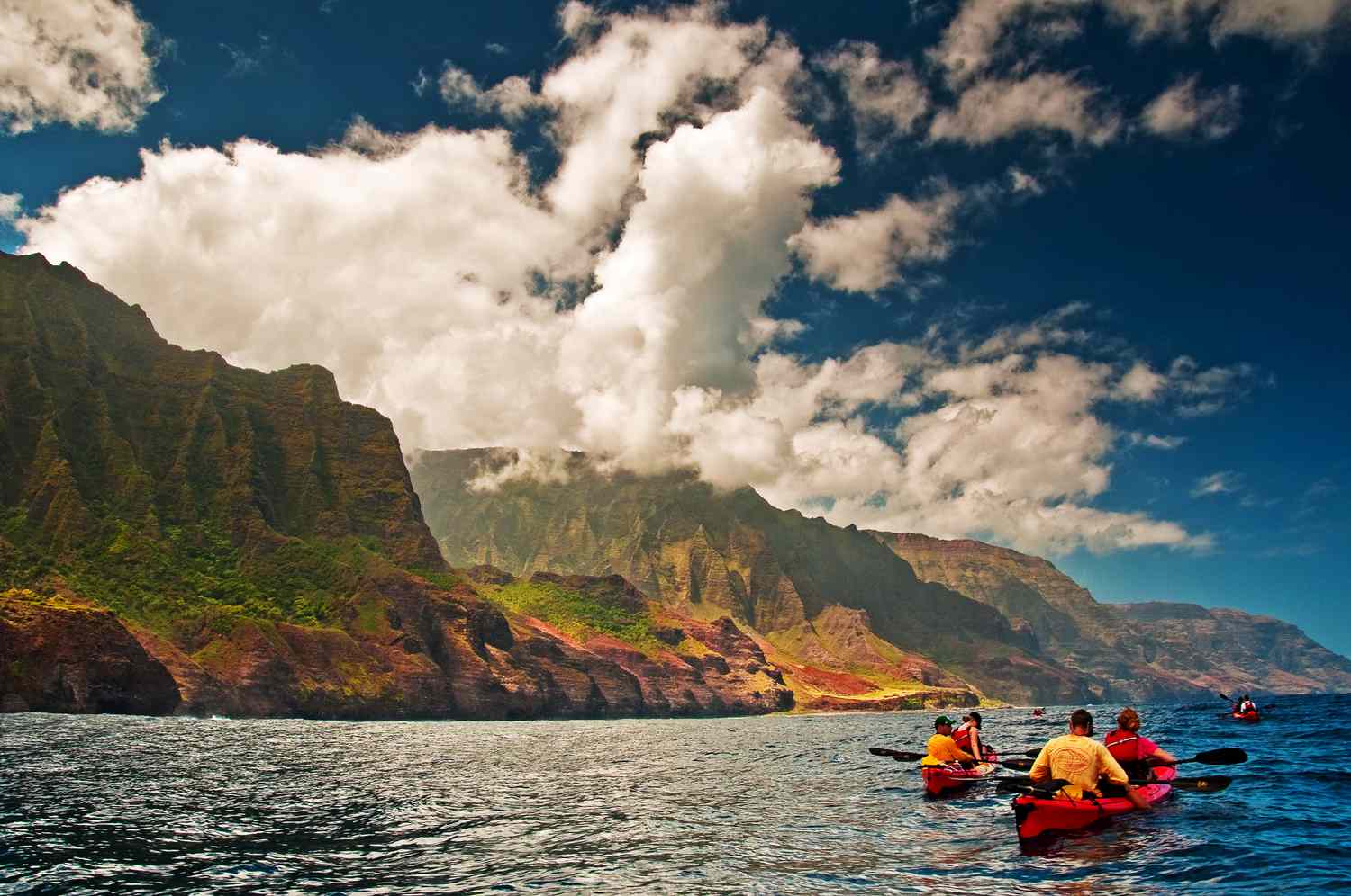 two pairs of kayakers in the waters off of Na Pali Coast, Kauai next to flat-topped rocks covered in green plants, blue sky, and white, billowy clouds