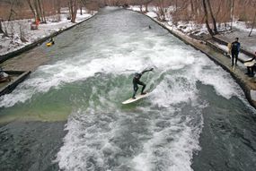 Surfer in the middle of the Eisbach surfing in winter with snow on both sides of the river and a forest of trees with brown leaves
