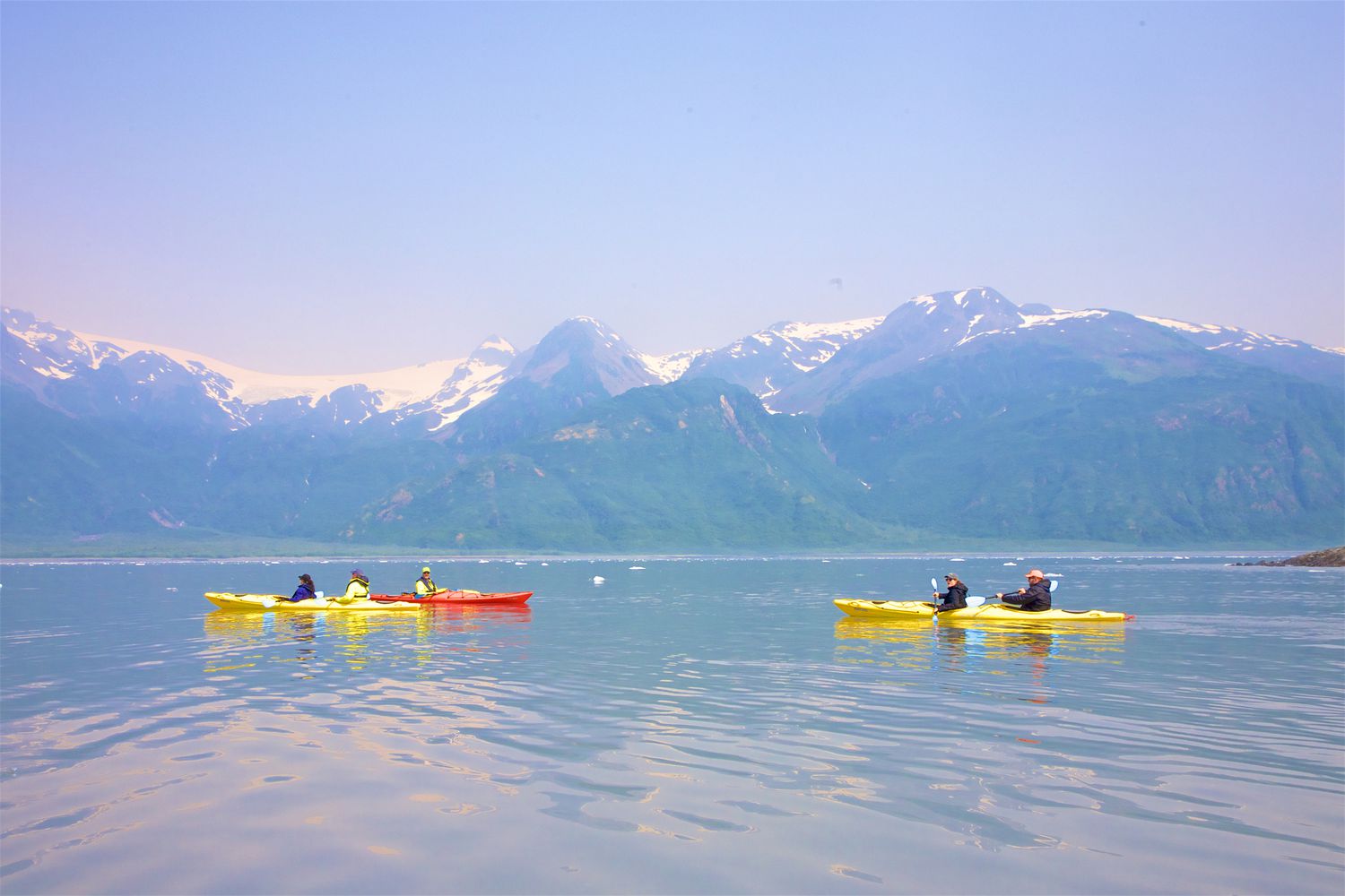 kayakers in yellow and red kayaks in the water off of Kenai Fjords National Park with snow-capped mountains and blue sky in the distance