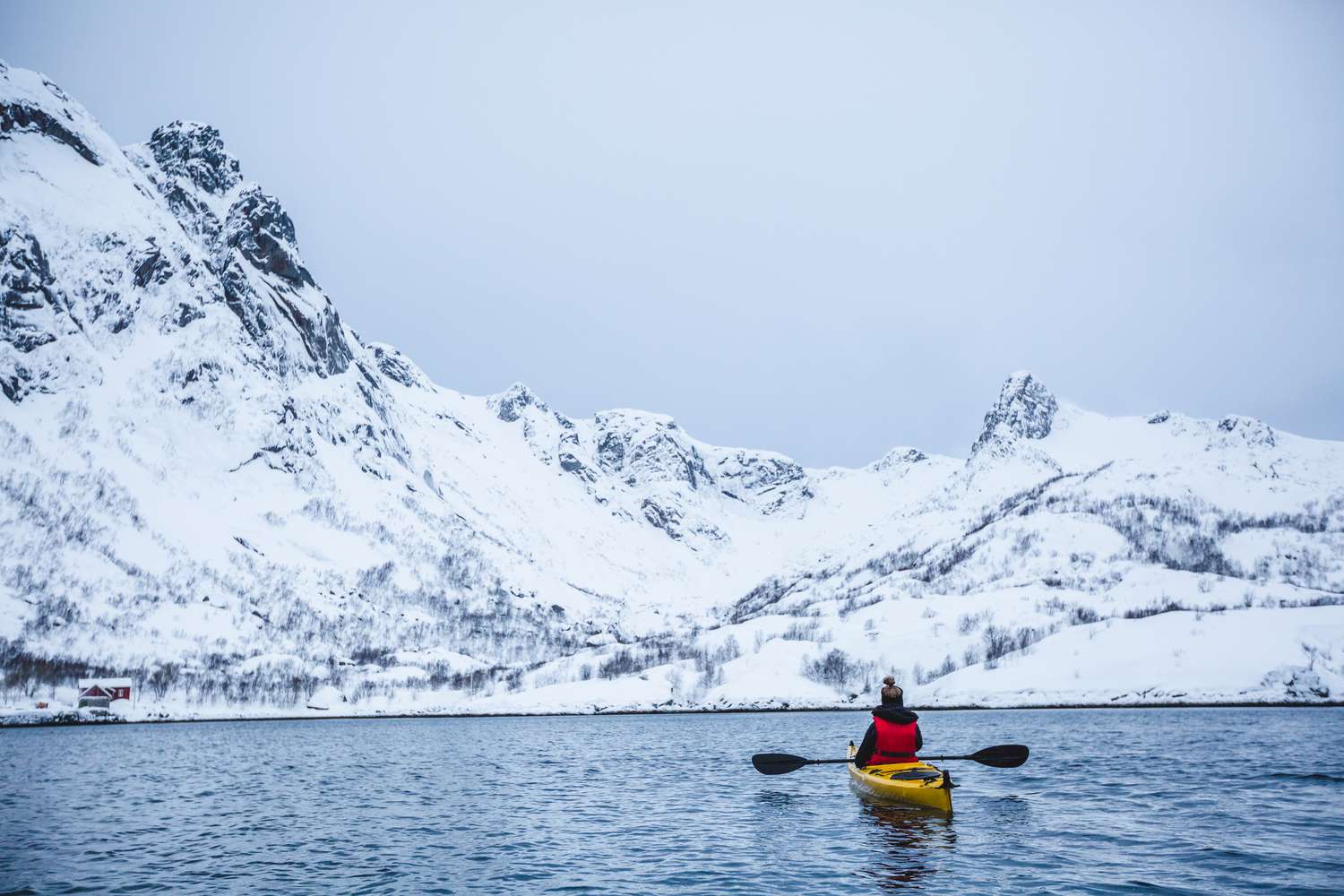 A single kayaker in a yellow kayak with paddles outstretched facing a snow covered fjord in Norway
