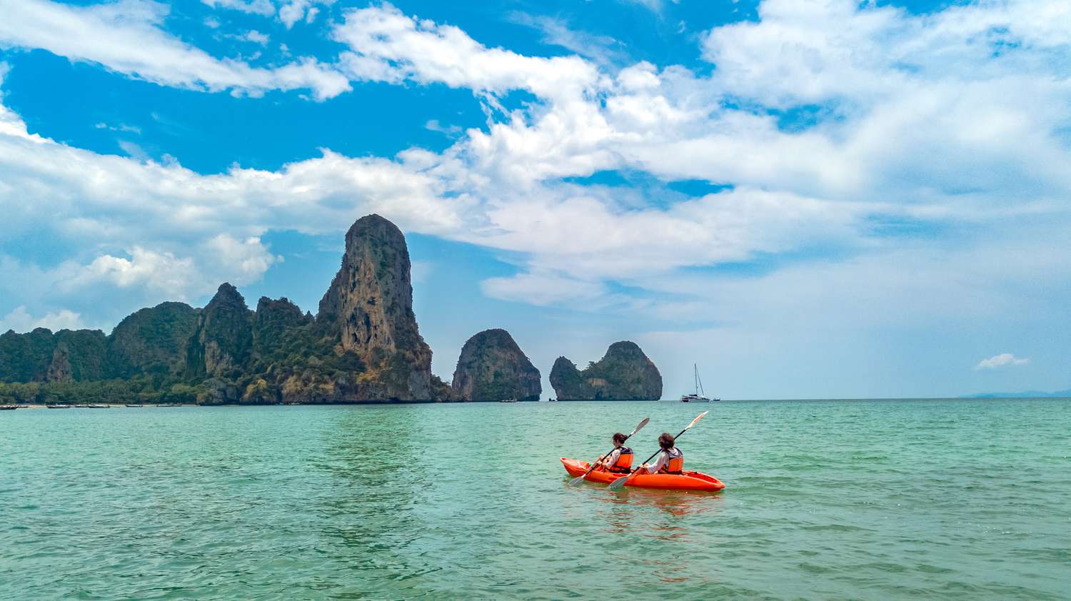 Two people in a red kayak paddling in blue-green water toward a rock formation in Krabi, Thailand.