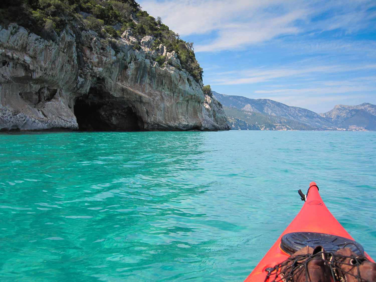 front of a red kayak floating in the blue-green waters heading toward the coves and caves of Gulf of Orosei on Eastern Sardinian coast with blue skies and light, white clouds above