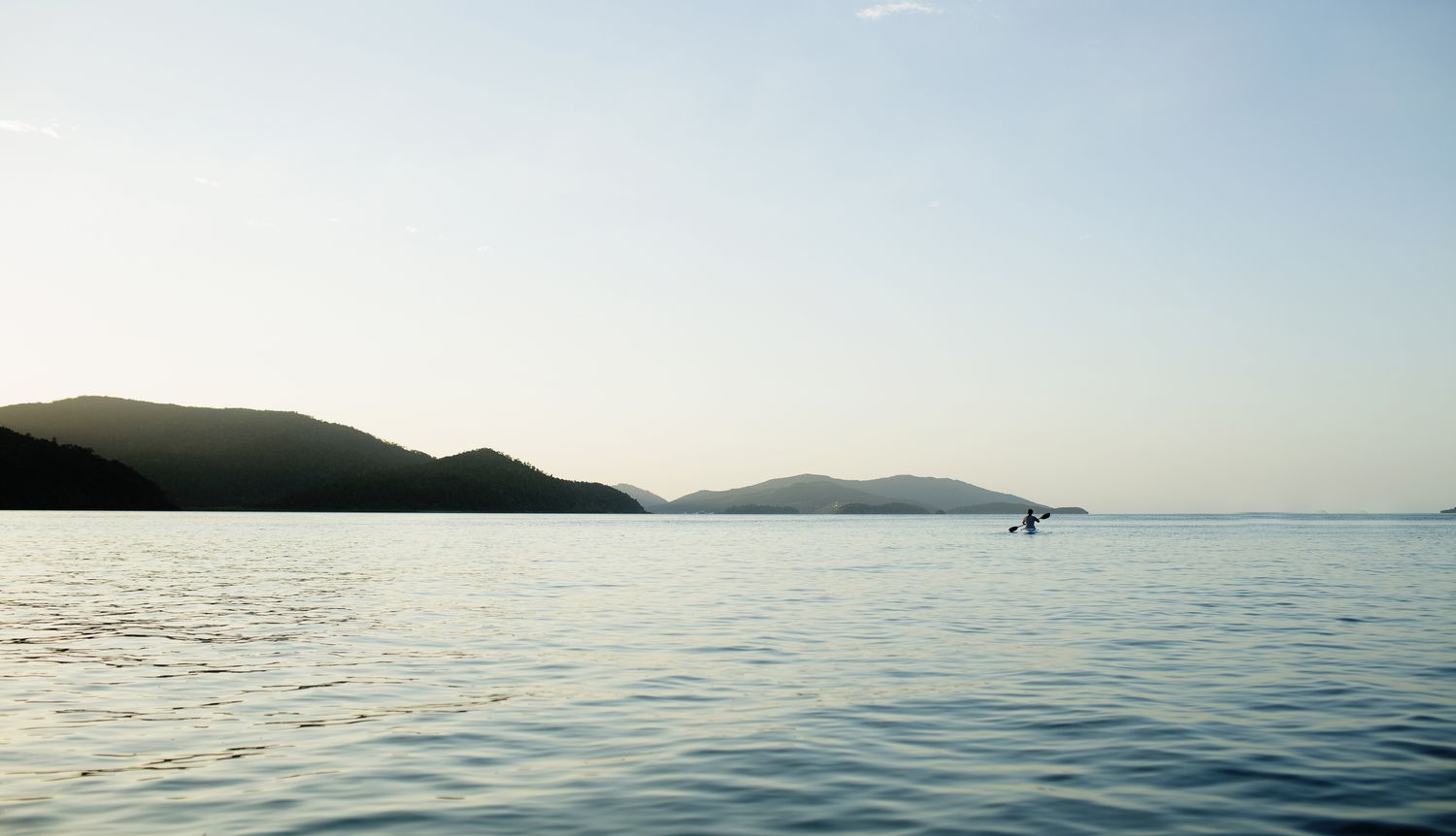 Lone kayaker in the blue waters off the Whitsunday Islands off the coast of Queensland with clear blue sky and hills in the distance