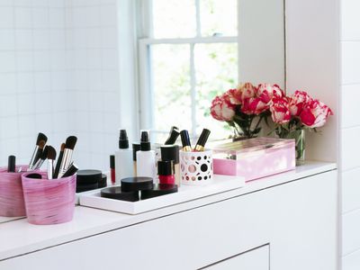 Makeup organized in pink containers on a bathroom ledge with a vase of pink tulips in the corner