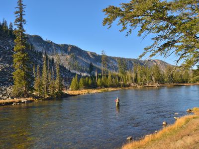 Man fly fishing in the middle of a stream surrounded by mountains and tall green trees on the Yellowstone River at Yellowstone National Park.