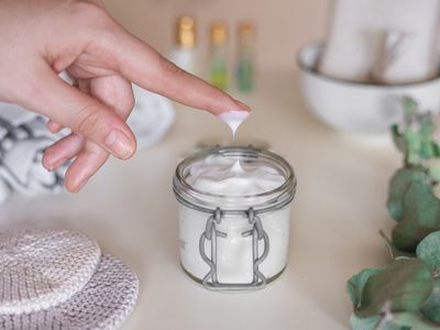 hand dips finger into glass jar of homemade lotion made with oils and beeswax