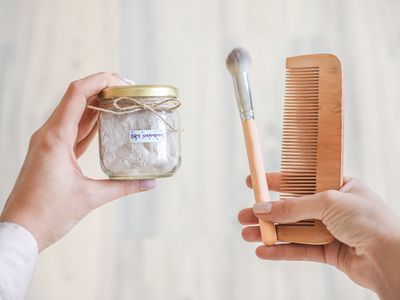 hand holds diy homemade dry shampoo in glass jar and other hand holds comb and brush