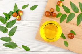 Jojoba oil in a bowl, a sprig of jojoba and fruits on a wooden table, top view