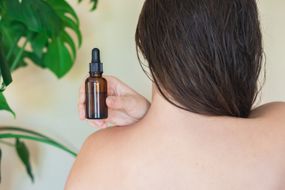 woman's bare back with hair swept aside while she displays glass bottle of moringa oil for skin