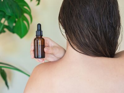 woman's bare back with hair swept aside while she displays glass bottle of moringa oil for skin