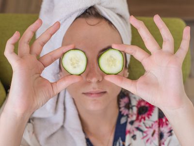 women wearing towel head wrap and robe holds up cucumber slices in front of eyes