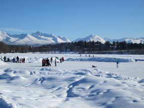 Mounds of snow give way to a frozen lake with dozens of ice skaters on it as snowy mountains loom in the background on a blue-sky afternoon